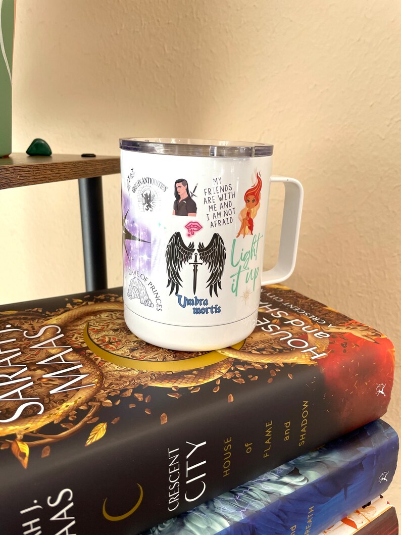 Puede incluir: Taza de viaje blanca con un gr&aacute;fico en blanco y negro de alas y una espada, con el texto "Umbra mortis" y "Light it up". La taza tambi&eacute;n tiene otros gr&aacute;ficos, incluyendo un personaje con una espada, un personaje con una corona y un personaje con una llama. La taza est&aacute; encima de una pila de libros con el t&iacute;tulo "Crescent City" y "House of Flame and Shadow".