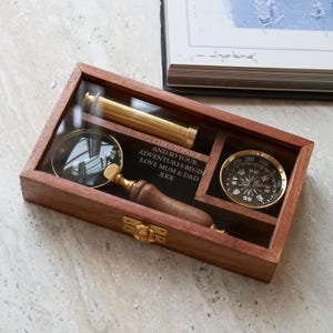 Puede incluir: Una caja rectangular de madera con tapa de cristal que contiene un telescopio de latón, una lupa con mango de madera y una brújula de latón. La tapa de cristal tiene grabado el texto: CHRISTOPHER, AND SO YOUR ADVENTURES BEGIN LOVE MUM & DAD XXX.