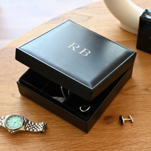 May include: A square black jewellery box with a hinged lid featuring the silver embossed letters RB. The interior is lined with black fabric and contains compartments holding a silver chain and a single black cufflink, with a watch nearby.