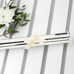 Può includere: Un portadiploma argentato con un nastro color crema. Il testo "Congratulations! Dr. Hannah Roberts" è inciso sul tubo. Fiori bianchi e fogliame verde sullo sfondo.