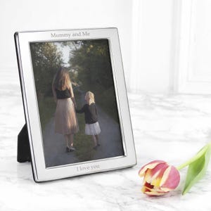 May include: A silver photo frame displaying a picture of a mother and child. The frame has the words "Mummy and Me" and "I love you" engraved on it. A pink and yellow tulip is placed in the foreground, next to the frame.