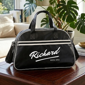 May include: A black duffel bag with white piping and two top handles. The front features the white printed text Richard in a cursive script, with the text ESTD 1994 printed in a smaller sans-serif font underneath the underlined name.