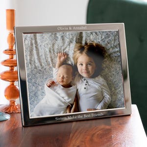 Op de afbeelding: Een rechthoekige fotolijst van zilverkleurig metaal met de gegraveerde tekst Olivia & Annabel aan de bovenkant en Sisters Make the Best Friends aan de onderkant. De lijst bevat een foto van twee kinderen op een bloemrijke stof.