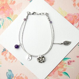 May include: A delicate silver bracelet with a floral and leaf charm, a small disc with the letter "A", and two purple amethyst beads. The bracelet is displayed on a white surface with a floral background.