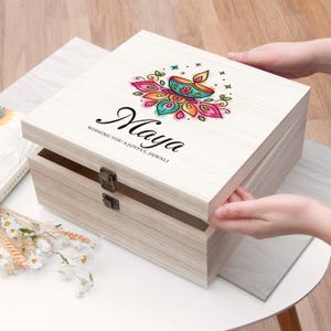 May include: A light-coloured wooden keepsake box with a hinged lid featuring a colourful diya lamp illustration, the name Maya, and the text WISHING YOU A JOYFUL DIWALI. The box has two metal latches on the front and a smooth, light wood grain finish.