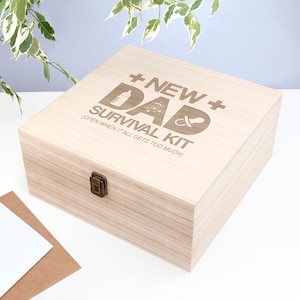 May include: A light wood box with a hinged lid and a brass latch. The box is engraved with the text "New Dad Survival Kit (Open when it all gets too much)" and includes icons of a baby bottle, a rattle, and a pacifier.