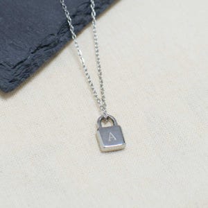 Puede incluir: Collar plateado con un pequeño colgante de candado cuadrado. El colgante tiene grabada la letra "A". La cadena es fina y el collar se muestra sobre un fondo neutro.