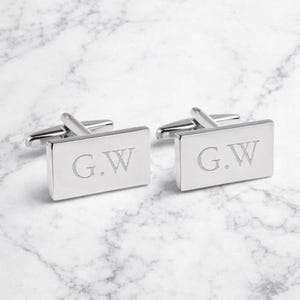 Puede incluir: Un par de gemelos rectangulares plateados con las iniciales "G.W." grabadas. Se exhiben sobre una superficie de mármol blanco. Un accesorio elegante y atemporal.