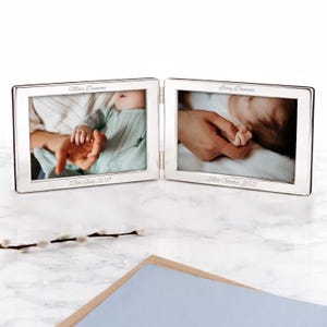 May include: A silver-colored, hinged double photo frame. Each frame holds a photograph of a baby's hand in an adult's hand. The names Miles Duncan and Lacey Duncan are engraved above the photos, with dates below.