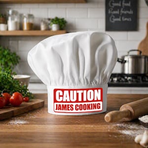 Puede incluir: Un gorro de chef blanco con el texto "CAUTION JAMES COOKING" en un rectángulo rojo. El gorro está sobre una superficie de madera con un rodillo, tomates y otros utensilios de cocina en el fondo. El gorro es de tela suave y plisada.