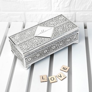 May include: A rectangular silver-toned metal keepsake box featuring an intricate embossed floral pattern across the lid and sides. A smooth, diamond-shaped central plaque on the lid is engraved with the cursive name ella.