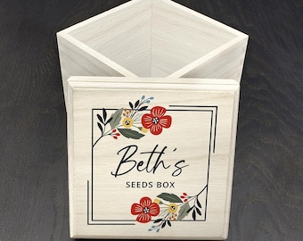 May include: A square, light-coloured wooden box with an open top and a matching lid. The lid features a black rectangular border, floral illustrations in red, yellow, and dark green, and the text "Beth's SEEDS BOX" printed in black.
