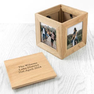 May include: A wooden photo cube with family photos displayed on three sides. The cube has a removable lid with engraved text: "The Wilsons Lake District 31st April 2024". The cube is made of light-colored wood and sits on a white surface.