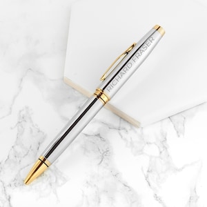 May include: A silver ballpoint pen with gold-colored accents and a gold-colored clip. The barrel of the pen is engraved with the name RICHARD FRASER in capital letters. The pen features a polished metallic finish and a gold-colored tip.