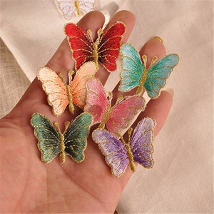 May include: Assortment of colorful embroidered butterfly iron-on patches. The patches feature a variety of colors, including red, pink, green, blue, and purple. Each butterfly has a gold outline and is approximately 2 inches in size.