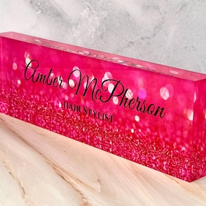 May include: Pink acrylic block with a glittery background and the text "Amber McPherson Hair Stylist" in black.