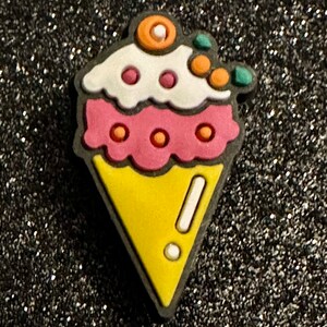 May include: A colorful ice cream cone charm with a yellow cone, pink ice cream, and white frosting. The charm has orange and green accents and a black outline.