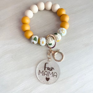 May include: A white and yellow beaded keychain with a floral pattern. The keychain has a metal ring and a clear charm with the text "Fur Mama" and a small heart.