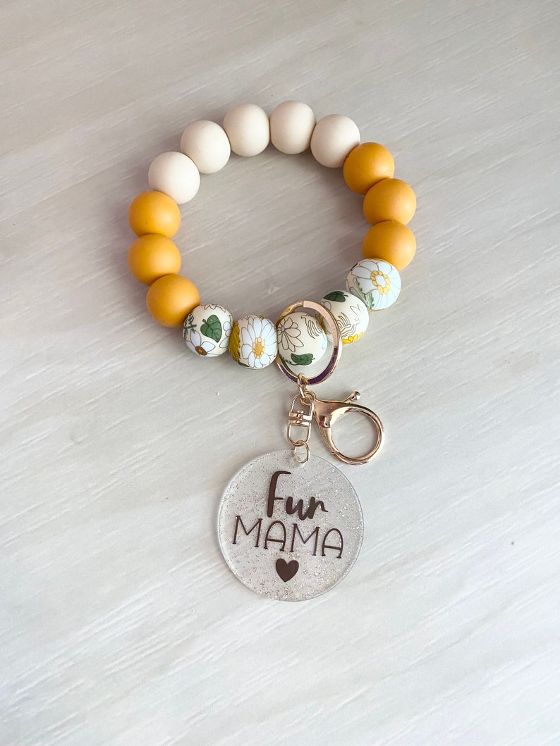 May include: A white and yellow beaded keychain with a floral pattern. The keychain has a metal ring and a clear charm with the text "Fur Mama" and a small heart.