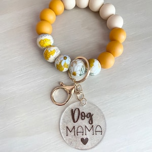 May include: A yellow and white beaded keychain with a floral pattern. The keychain has a gold metal clasp and a clear round charm with the text "Dog Mama" and a small heart.