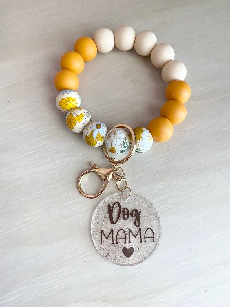 May include: A yellow and white beaded keychain with a floral pattern. The keychain has a gold metal clasp and a clear round charm with the text "Dog Mama" and a small heart.