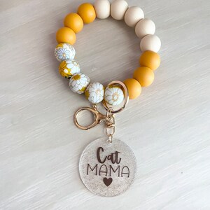 May include: A yellow and white beaded keychain with a clear charm that says "Cat Mama" with a small heart.