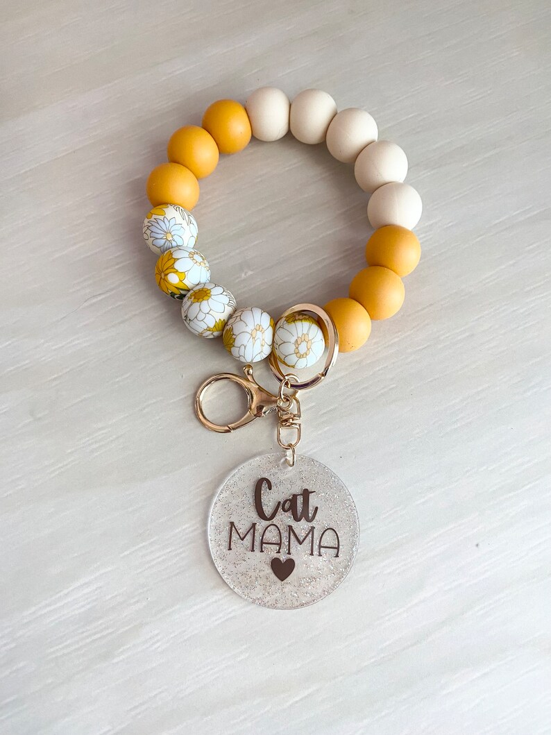 May include: A yellow and white beaded keychain with a clear charm that says "Cat Mama" with a small heart.