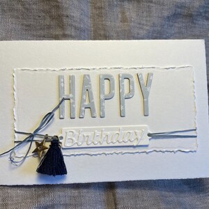 Pode incluir: Um cartão de aniversário feito à mão com a palavra "HAPPY" em letras cinzentas. A palavra "Birthday" está abaixo. Uma borla azul marinho e um berloque de estrela dourada são presos com barbante. O cartão é branco com uma borda texturizada.