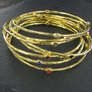 May include: A stack of gold-colored bangles with a textured surface. Each bangle is adorned with small, colorful gemstones, including red, blue, and clear stones. The bangles are stacked on a dark surface.