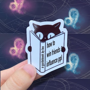 May include: A white sticker featuring a black cat reading a book titled "how to win friends & influence ppl" with the text "a guidebook by leo". The background includes a dark blue celestial design with Leo zodiac symbols.