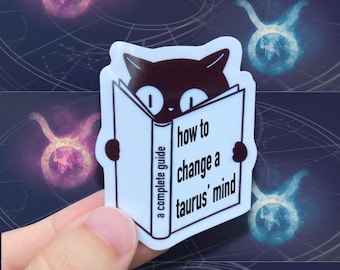 May include: A white sticker featuring a black cat reading a book titled "how to change a taurus' mind: a complete guide." The background includes a dark blue celestial design with Taurus zodiac symbols in pink and blue.