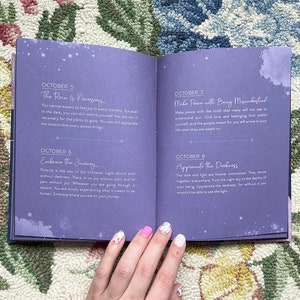 May include: Open book with purple pages featuring daily inspirational quotes for October. The text includes phrases like "The Rain Is Necessary" and "Embrace the Journey." The book is open on a floral patterned rug.