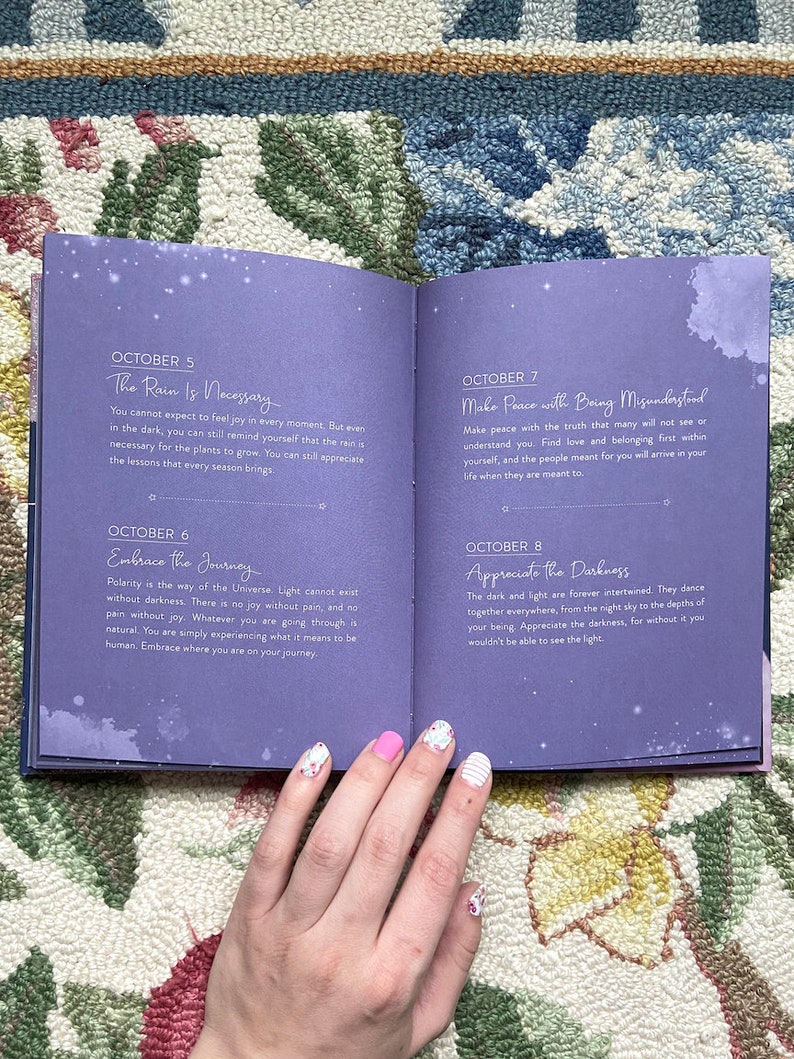 May include: Open book with purple pages featuring daily inspirational quotes for October. The text includes phrases like "The Rain Is Necessary" and "Embrace the Journey." The book is open on a floral patterned rug.