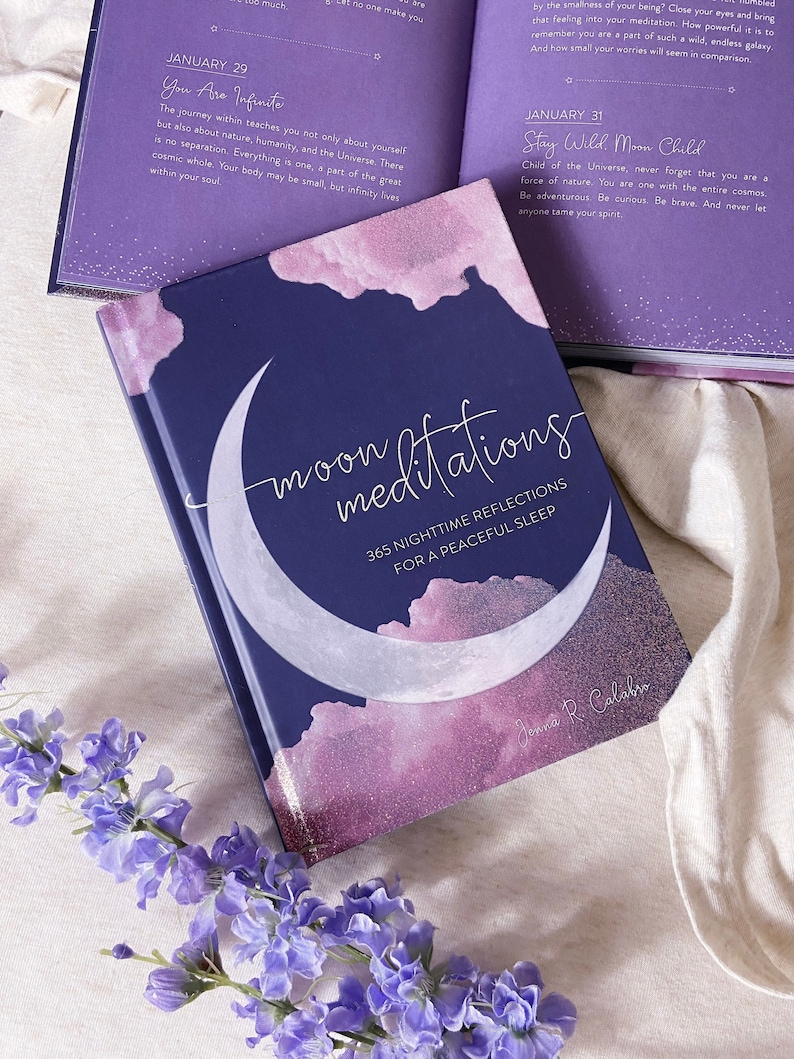 May include: A hardcover book titled "moon meditations" with a crescent moon design and the text "365 Nighttime Reflections for a Peaceful Sleep." The book is navy blue with pink and purple accents. Open pages of a journal are visible.