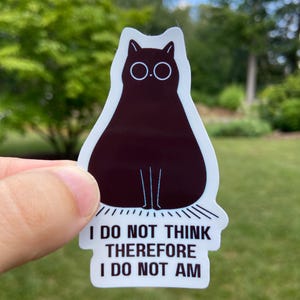 May include: A sticker featuring a dark brown cat silhouette with white eyes and the text "I DO NOT THINK THEREFORE I DO NOT AM". The cat sticker is held up against a green background of trees and grass.