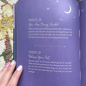 May include: Open journal pages with text about guidance and releasing the past. The left page has text that reads "August 28 You Are Being Guided" and "August 29 Release Your Past". The pages are purple with white text and star and moon illustrations.