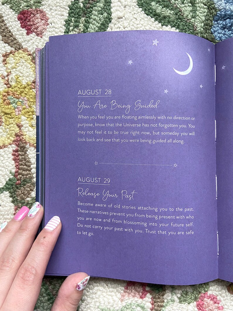 May include: Open journal pages with text about guidance and releasing the past. The left page has text that reads "August 28 You Are Being Guided" and "August 29 Release Your Past". The pages are purple with white text and star and moon illustrations.
