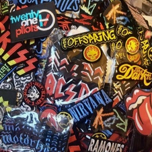 May include: A selection of band patches featuring logos from bands like Twenty One Pilots, KISS, Nirvana, and The Offspring. The patches are in various shapes and sizes, with bright colours and detailed designs. These patches are likely for clothing or accessories.