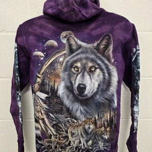 Peut inclure: Un sweat à capuche violet avec un motif de loup détaillé. Le devant présente le visage d'un grand loup, tandis que le dos affiche une meute de loups dans un cadre forestier, avec un attrape-rêves et des éléments célestes.
