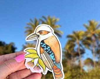 May include: A die-cut sticker featuring a colorful illustration of a kookaburra bird perched on a branch with yellow leaves and pink flowers. The bird has a white head, brown eye, tan back with white patterns, blue wing feathers, and a striped tail.