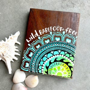 May include: A wooden sign featuring a teal, green and yellow mandala design with the text "wild barefoot + free" painted in white.