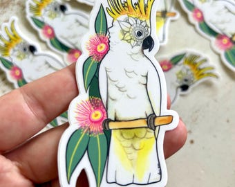 May include: A die-cut sticker featuring a white cockatoo with a yellow crest and yellow tail feathers, perched on a brown branch. The bird is decorated with a black and white mandala pattern on its face and is surrounded by green leaves and pink flowers.