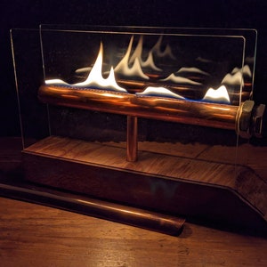 Op de afbeelding: Een tafelhaard met een koperen pijp en een glazen omhulling. De vlammen zijn zichtbaar door het glas.