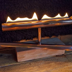 Op de afbeelding: Een koperen tafelhaard met een houten basis. De haard heeft drie vlammen die van boven branden.