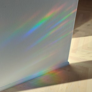 Op de afbeelding: Een witte muur met een regenboogprisma lichtreflectie. Het licht werpt een schaduw op de lichte houten vloer.