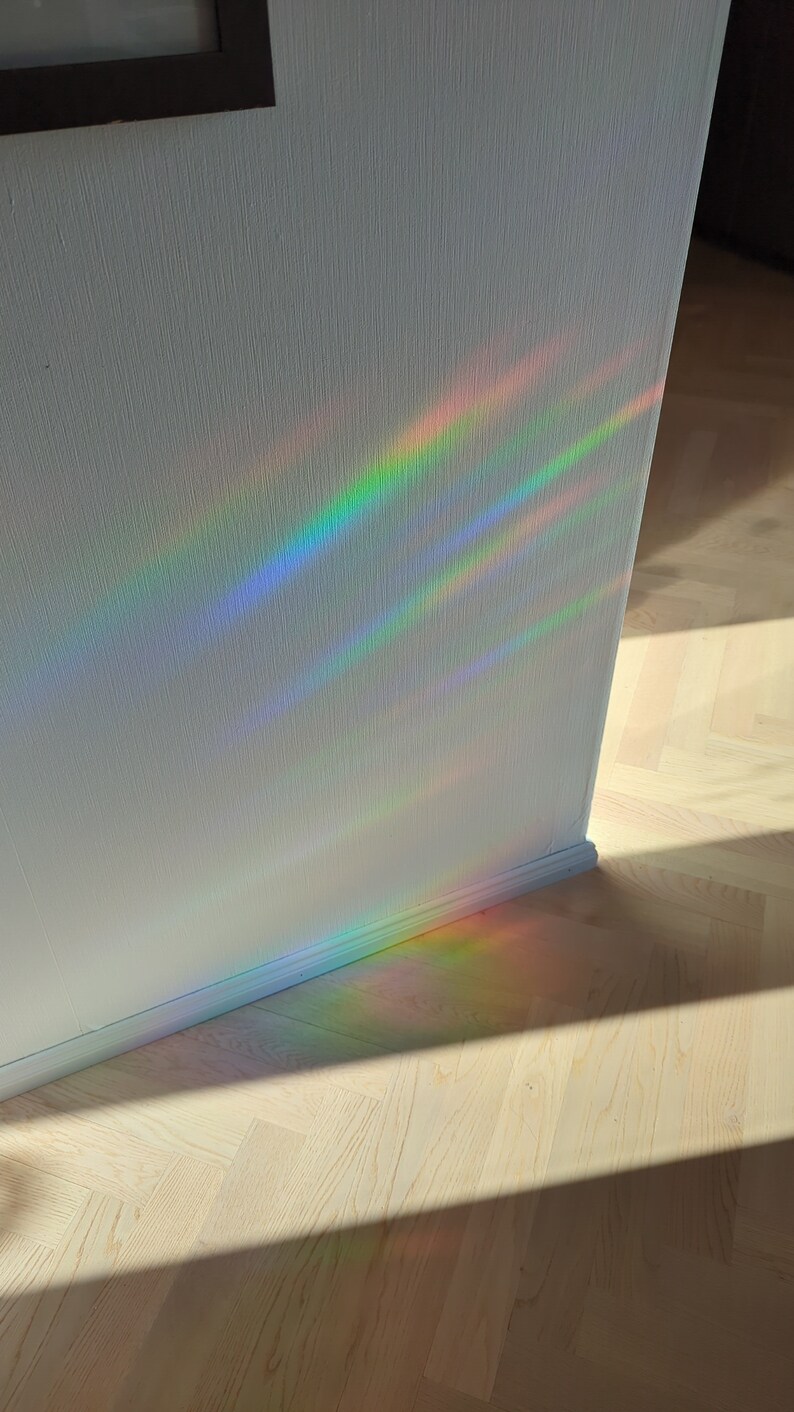 Op de afbeelding: Een witte muur met een regenboogprisma lichtreflectie. Het licht werpt een schaduw op de lichte houten vloer.
