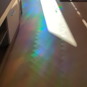 Puede incluir: Un rayo de sol brilla a trav&eacute;s de una ventana, creando un efecto de prisma de arco&iacute;ris en el suelo. La luz crea un patr&oacute;n colorido y abstracto en el suelo marr&oacute;n claro.