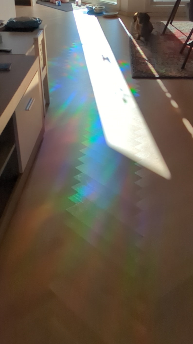 Puede incluir: Un rayo de sol brilla a trav&eacute;s de una ventana, creando un efecto de prisma de arco&iacute;ris en el suelo. La luz crea un patr&oacute;n colorido y abstracto en el suelo marr&oacute;n claro.