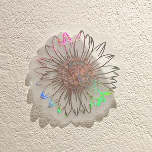 Puede incluir: Una pegatina hologr&aacute;fica con un contorno en blanco y negro de un girasol. La pegatina tiene un acabado iridiscente arco&iacute;ris.