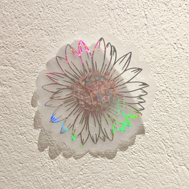 Puede incluir: Una pegatina hologr&aacute;fica con un contorno en blanco y negro de un girasol. La pegatina tiene un acabado iridiscente arco&iacute;ris.